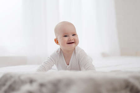 Portrait of a cute beautiful baby with gray eyes in white pajamas on the bed against the window....