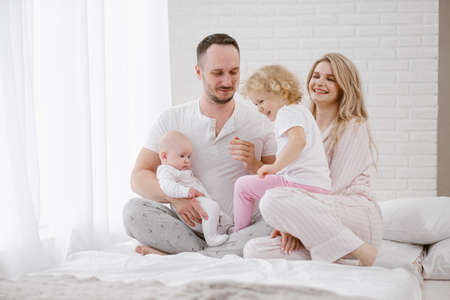 A young Caucasian family with two children daughters in a white home interior. Home clothes. People are sitting on the bed in the bedroom.の写真素材