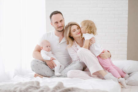 A young Caucasian family with two children daughters in a white home interior. Home clothes. People are sitting on the bed in the bedroom.の写真素材