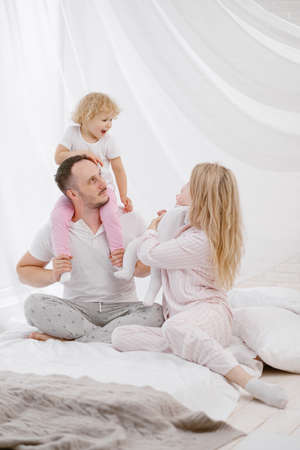 A young Caucasian family with two children daughters in a white home interior. Home clothes. People are sitting on the bed in the bedroom.の写真素材