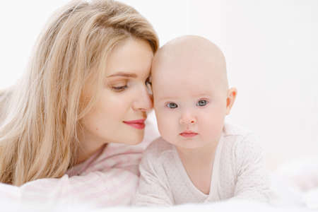 Young beautiful Caucasian blonde mom lies on the bed with her baby daughter. White light home interior. Love and care.の写真素材