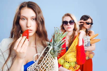 Three young fashionable females housewives in red and blue swimsuits and trench coats on naked body. Shopper strip bags with food in hands. Shopping, purchasing and consumerism. Beautiful long legs.の写真素材
