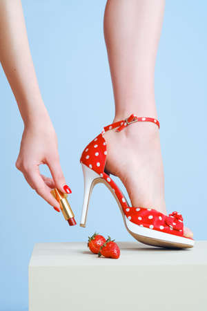 Lipstick in hand and red high-heeled shoes close-up on a blue background. Advertising cosmetics and shoes.の写真素材