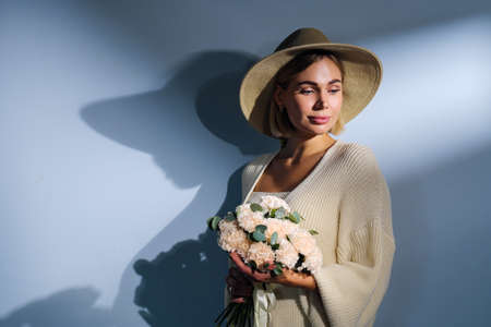 A young beautiful blonde caucasian woman with a short haircut in a beige suit and hat with bouquet of white flowers on blue background. Beauty portrait. The girl smiles, clean skin and white teeth.の写真素材