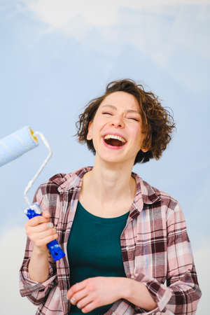 A young beautiful woman painter designer paints a white wall with blue paint with roller. Plaid shirt, short hair and jeans.の写真素材