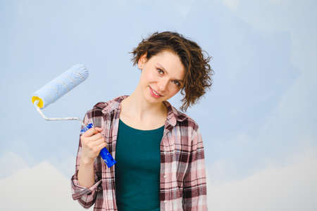 A young beautiful woman painter designer paints a white wall with blue paint with roller. Plaid...
