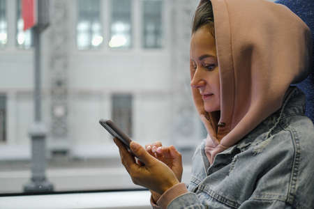 A beautiful young Caucasian woman in warm casual clothes sits by the window in a train or subway....
