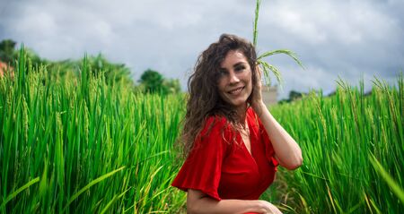 Portrait of young curly lovely woman wearing rice stalks. Romantic girl in red dress walks through rice fields. Happy preaty girl in the jungle near rice fields on the exotic island of Bali in Indonesia. Beautiful young lady in shine through dress touch rice stalks.の写真素材