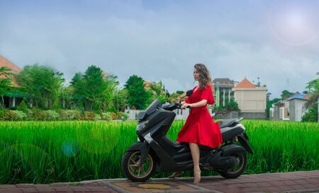 Young happy woman in red dress on a motorbike in the jungle drive around rice terrace of a tropical Bali island, Indonesia. Freedom concept. Rider Female on the scooter in motion. Woman riding on the motorbike on road near green rice. Fasion style on bike.の写真素材