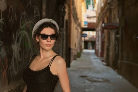 Beautiful style young woman wearing hat and in the city street in front of the old brick wall. Fashion tourist street concept. Outdoor fashion photo of young beautiful lady in hat and dress. Summer travel. Summer vibes.の写真素材