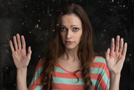 sad woman looking through the wet window. Dark backgroundの写真素材