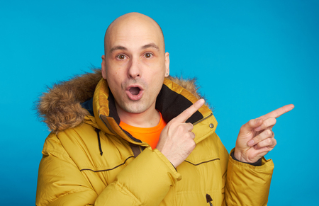 Amazed bald man wearing winter coat, looking with widely opened mouth at camera, pointing with forefinger at copy space for your textの写真素材