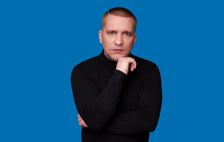 man thinking. Guy wearing black turtleneck jumper, looking at the camera, isolated on blue studio...