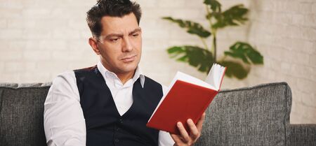Handsome man is reading a book while sitting on a sofa at home