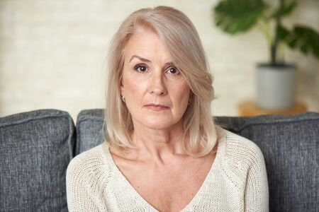 Sad senior blonde woman is sitting on a sofa at homeの写真素材