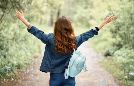 Back view of a pretty girl with brown curly hair in the park. Woman is raising hands wearing denim clothes and backpackの写真素材