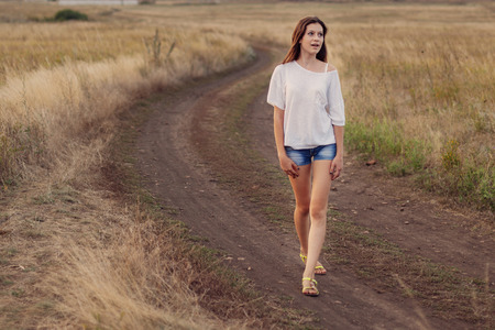 Young girl outdoors enjoying nature. She walking along the road in autumn field and looks happily. Selective focus, warm tinted.の写真素材
