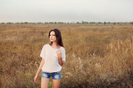 Young girl with long brown hair standing at the autumn meadow and looks thoughtfully left. Selective focus, warm tinted.の写真素材