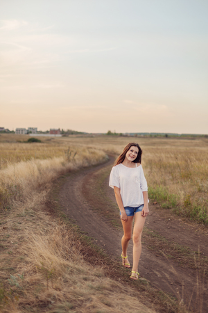 Young girl with long brown hair walking along the road in autumn field, smiling and looks happily. Selective focus, warm tinted.の写真素材