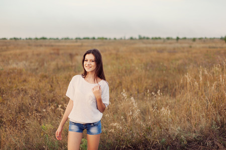 Young girl with long brown hair standing at the autumn meadow and smiling. Selective focus, warm tinted.の写真素材