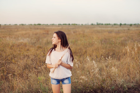 Young girl with long brown hair standing at the autumn meadow with spikelets in her hand, smiling and looks left. Selective focus, warm tinted.の写真素材