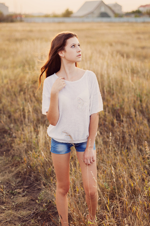 Young girl with long brown hair stay at the meadow and looks thoughtfully to right. Selective focus, warm tinted.の写真素材