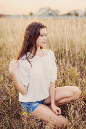 Young girl with long brown hair sitting in autumn field, smiling and looks happily. Selective focus, warm tinted.の写真素材