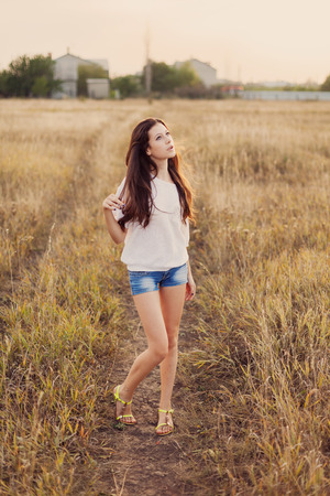 Young girl with long brown hair stay at the meadow and dreamily looks at the sky. She holds hand to head. Selective focus, warm tinted.の写真素材