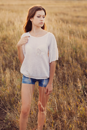 Young girl with long brown hair stay at the meadow and looks thoughtfully to right. Selective focus, warm tinted.の写真素材