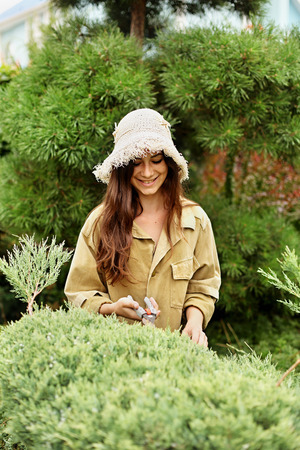 Girl gardener in working clothes and straw hat cuts garden scissors evergreen. She`s smiling.の写真素材