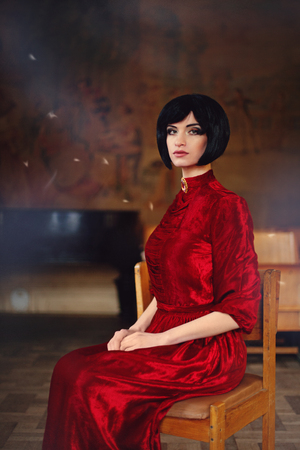 Portrait of lady brunette in the old castle. Beauty girl brunette with hairstyle and makeup in red vintage dress sitting at the old chair. Fairy tale, nostalgic mood. Warm tinted.の写真素材