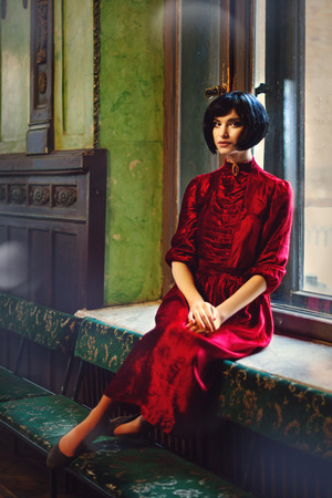 Portrait of lady brunette in the old castle. Beauty girl brunette with hairstyle and makeup in red vintage dress sitting at the window. Fairy tale, nostalgic mood. Warm tinted.の写真素材
