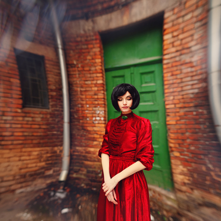 Young brunette girl in vintage romantic red dress near the vintage wooden door and brick wall. Fairy tale, nostalgic mood. Warm tinted. High resolution, Brenzier method image.の写真素材