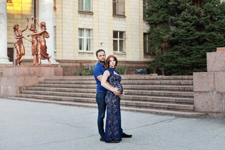Pregnant red hair woman in long blue dress and stylish man embrace outdoor in city.の写真素材