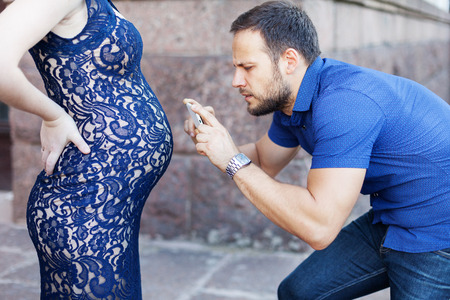 Funny image. Man photographing pregnant belly on the phone. Pregnancy in long dress. Future daddy in jeans and blue shirt.の写真素材