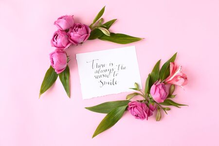 Card with calligraphic quote "There is always the reason to smile" on pink background. Rose buds and green leaves frame, flat lay, top view.の写真素材