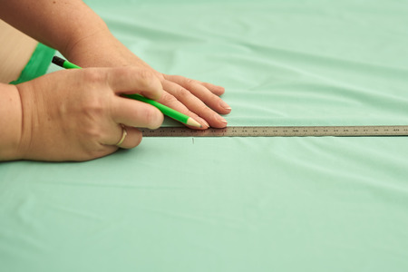 Woman cut out material green pencil for clothing. Green material lies on the table.