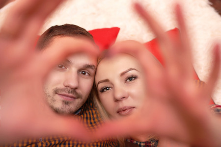 Girl with a guy lying on the bed and folded his fingers in the shape of a heart. A couple in love lies on red pillows.の写真素材