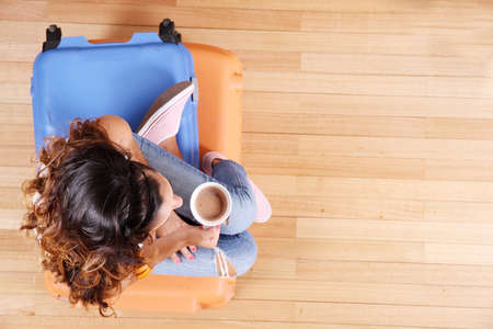 A young woman sitting on a stack of suitcases while drinking coffee and waiting for the departure to vacations の写真素材