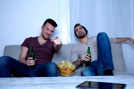 Two guys watching tv, drinking beer, eating crisps at homeの写真素材