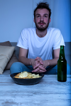 A young man watching a movie at nighttime with potato chips and beer at home in the living room.の写真素材