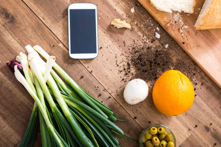 A smartphone lying in a natural kitchen with white bread, spices, olives and spring onions.の写真素材