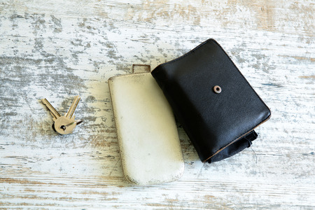 Keys, a wallet and a smartphone in a leather case on a wooden table.