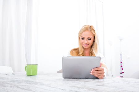 A beautiful young blond woman siting at the table having a coffee and using a tabletの写真素材
