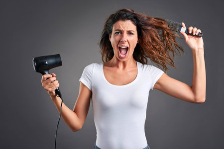 A beautiful young woman screaming while using a hairdryer and a hairbrush.