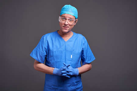 A middle aged male doctor standing in blue scrubs and rubber gloves and feeling confused