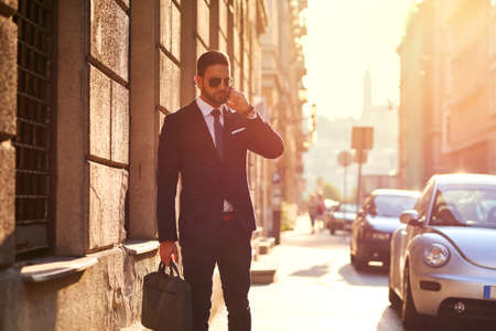 A handsome young businessman talking on his phone and walking on the street in the sunsetの写真素材