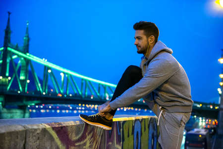 A handsome young sportsman ties his shoelace on the street at night on the riverside with a bridge...