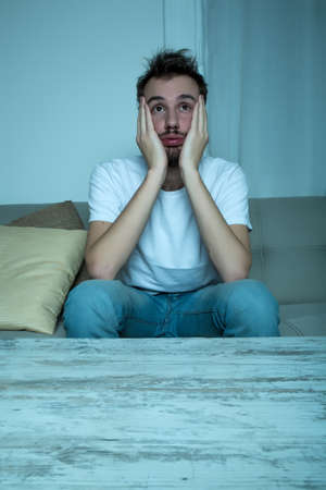 A young man sitting on the sofa in the living room is shocked by a movie on the TV at night.の写真素材