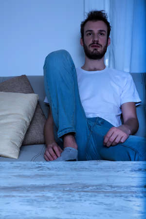 A young bored man sitting on the sofa in front of the TV at night while watching a movie.